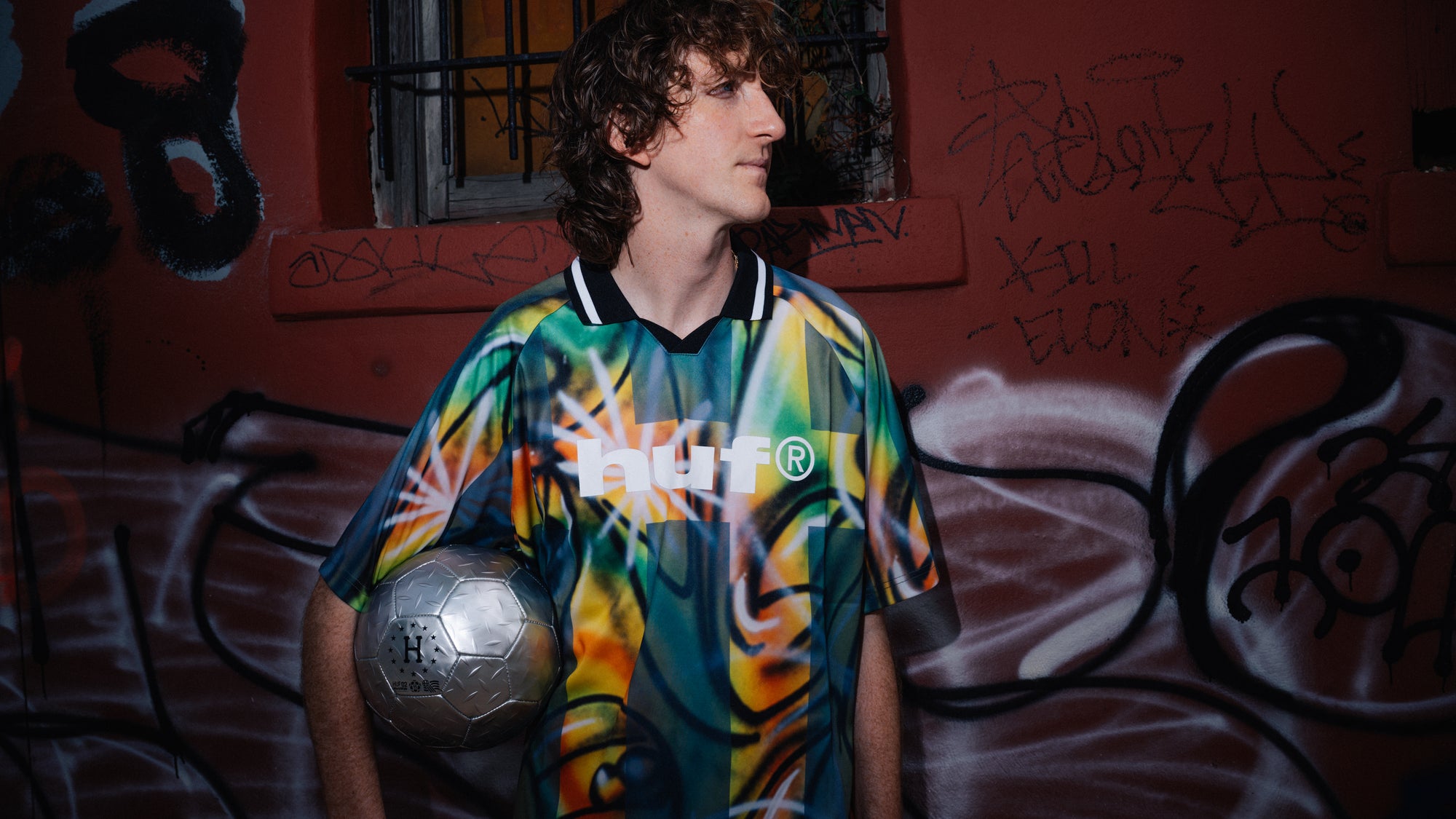 A man wearing a colorful retro soccer jersey with a 'HUF' logo, holding a soccer ball against a graffiti-covered red wall.